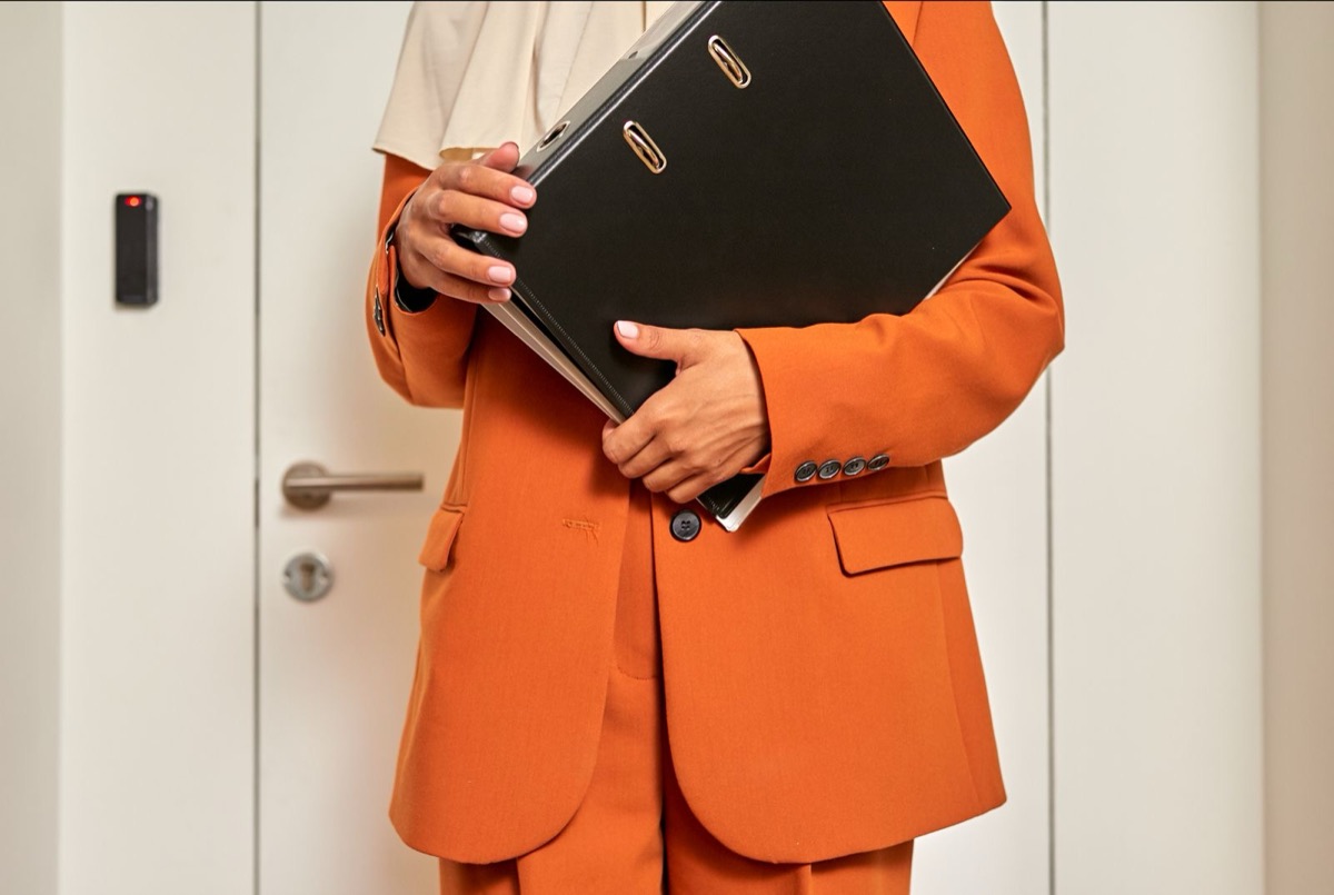 A person in an orange suit holding a closed black ring binder by a white door