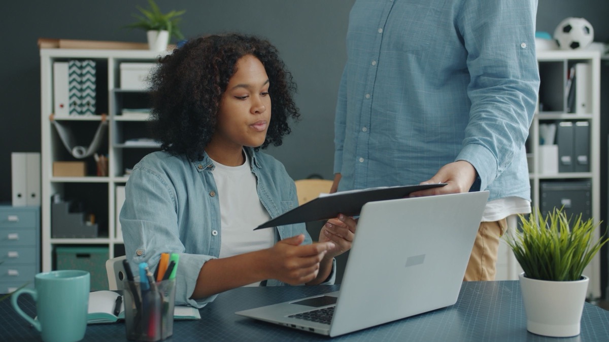 A woman seated at a laptop receiving a black folder from another person standing beside her