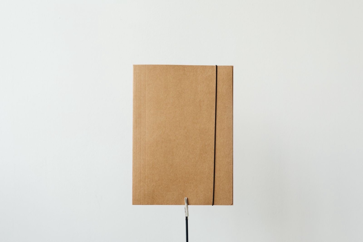 A closed kraft card folder with a black elastic band standing upright against a plain white wall