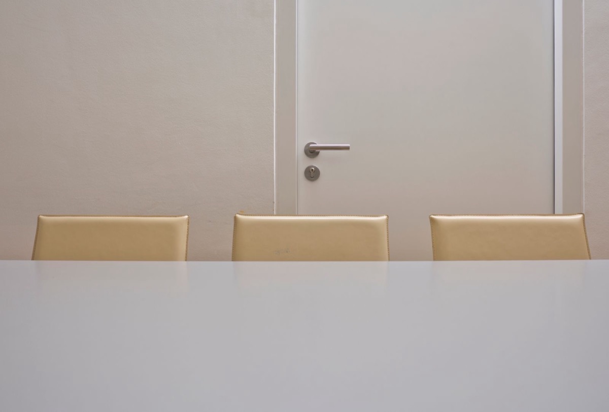 Three beige chairs behind a plain white table, facing a closed white door with a metal handle and lock