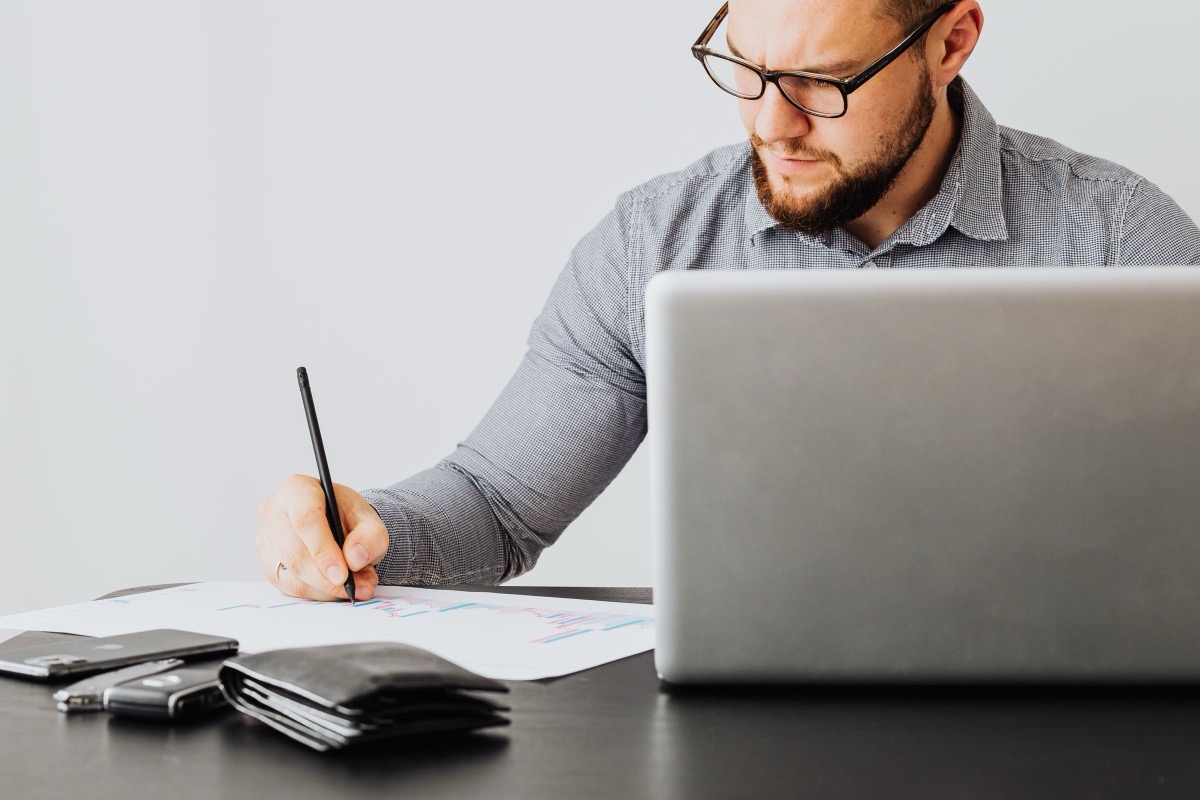 A man writing on a sheet of paper behind an open laptop, with a wallet and phone nearby