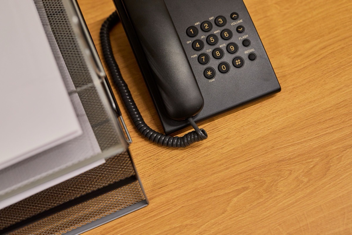 A black desk telephone beside a mesh paper tray holding sheets of paper on a wooden desk