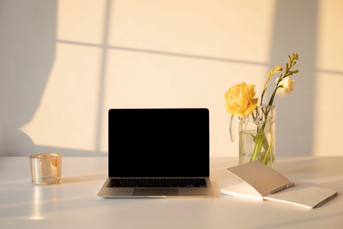 An open laptop with a blank screen, a vase of yellow flowers and a notebook on a white desk