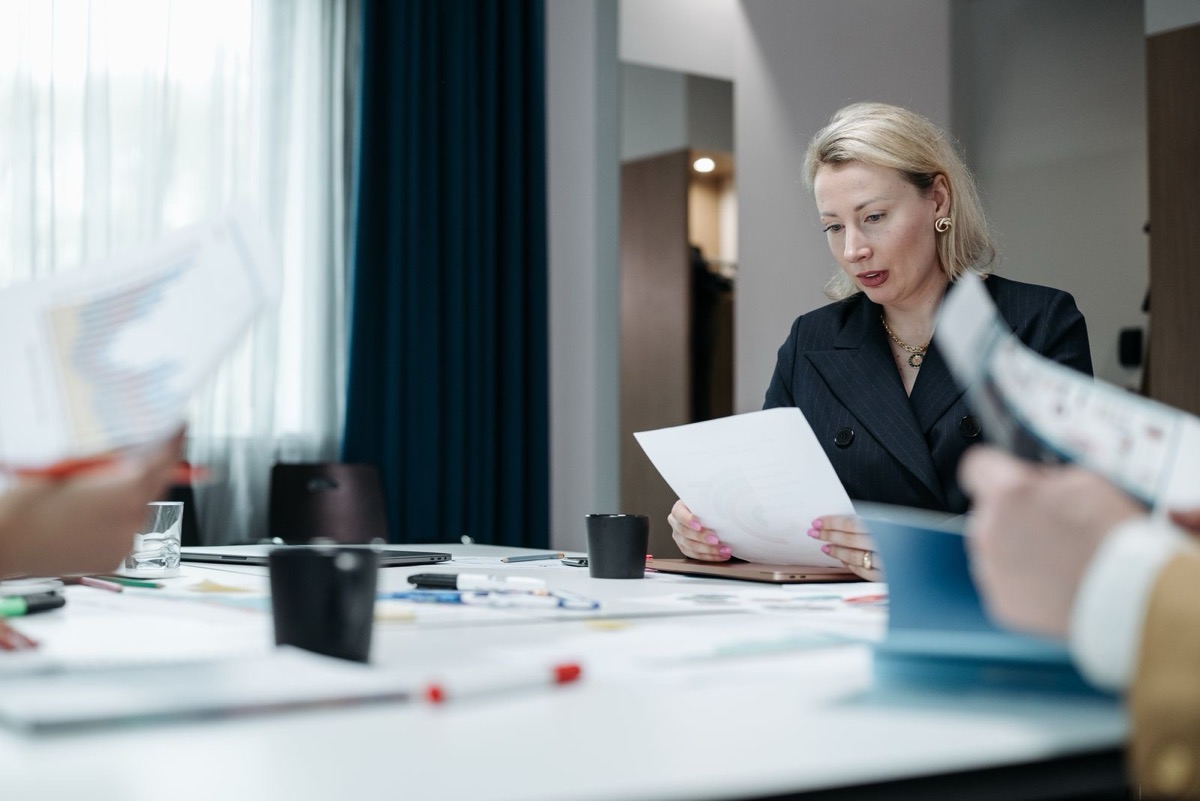 A woman in a dark blazer reading a sheet of paper at a meeting table with colleagues around her