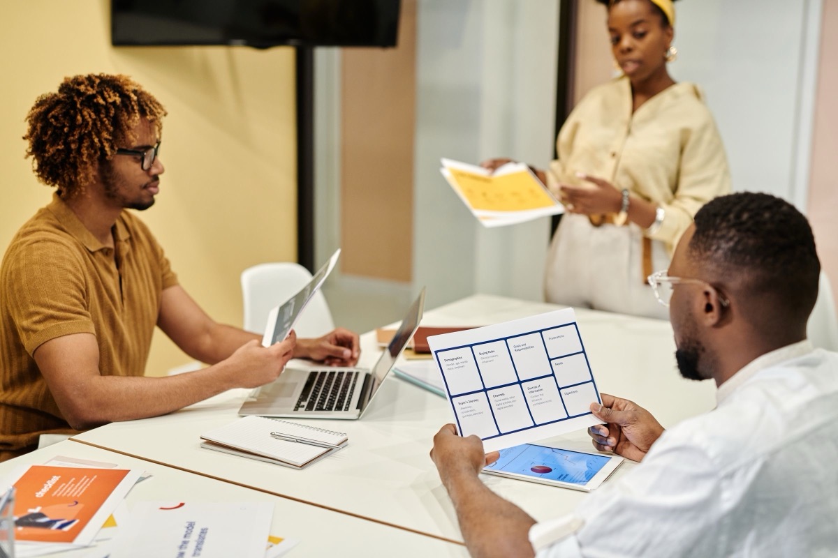 Three colleagues in a bright meeting room, two seated with a laptop and printed sheets, one standing with papers