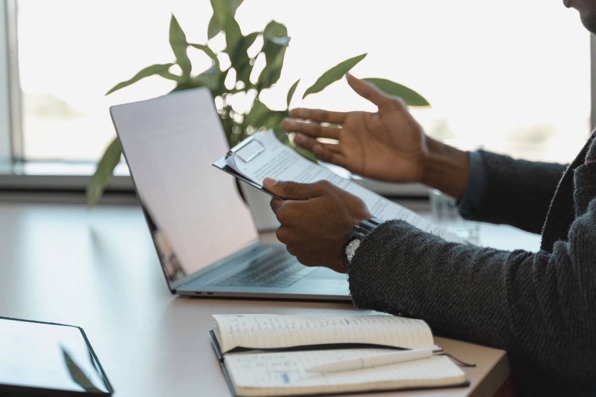 A person holding a printed document and gesturing beside an open laptop and a notebook on a desk