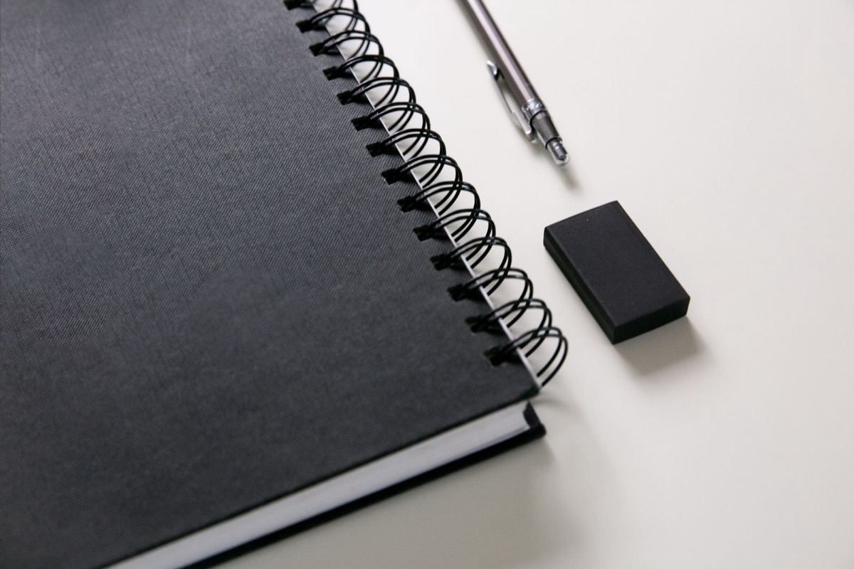 A closed black spiral notebook, a metal pen and a black eraser on a pale desk