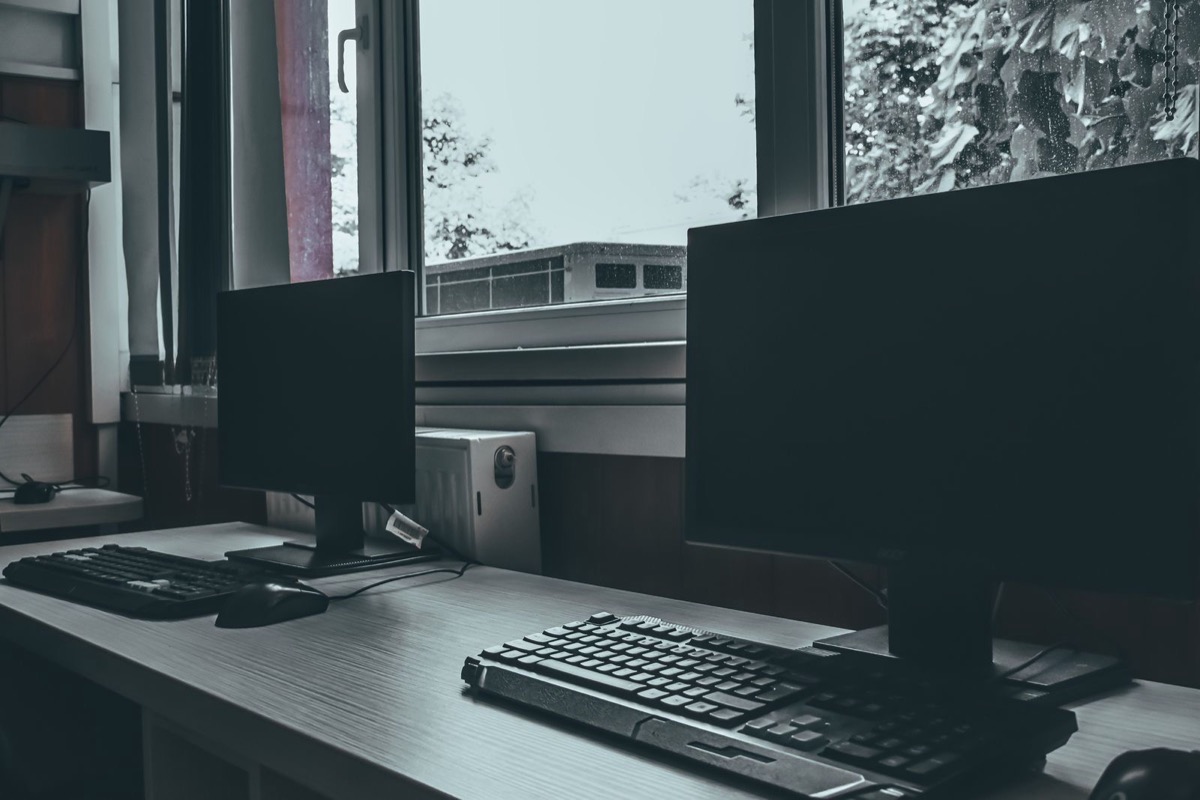 Two switched-off computer monitors with keyboards on a desk beside a window on a grey day