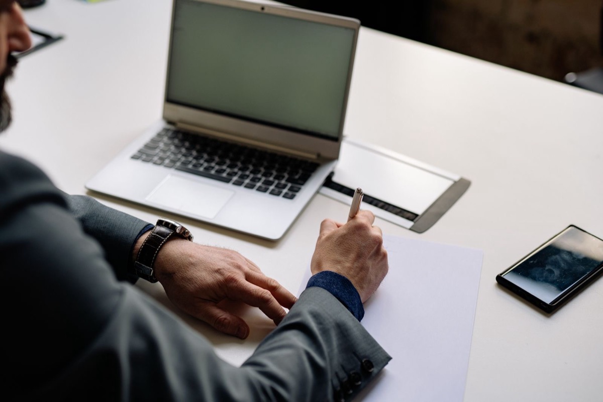 A hand holding a pen over a blank sheet beside an open laptop and a phone on a white desk
