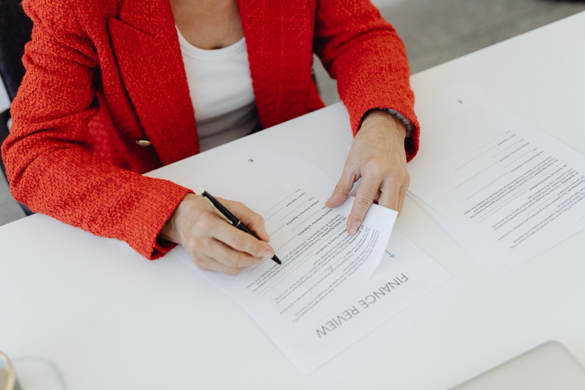 A person in a red jacket signing a printed document on a white desk, with another sheet beside it