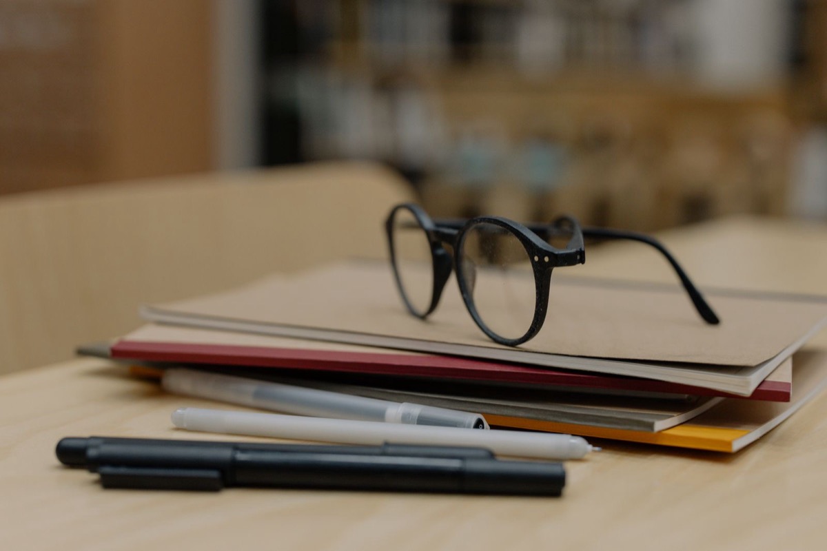 Stacked closed notebooks and folders with eyeglasses on top and pens in front, on a light wooden desk