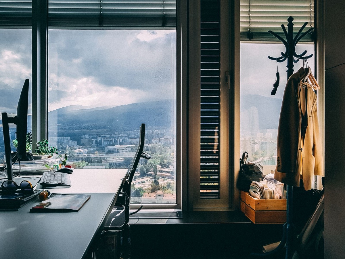 A desk with a monitor and keyboard beside tall windows overlooking a hilly city, with a coat on a stand