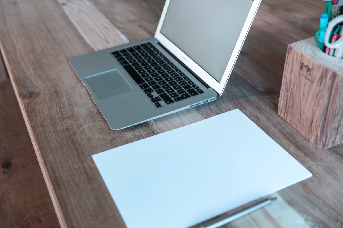 An open laptop, a blank sheet of paper and a pen on a wooden desk beside a pot of pens