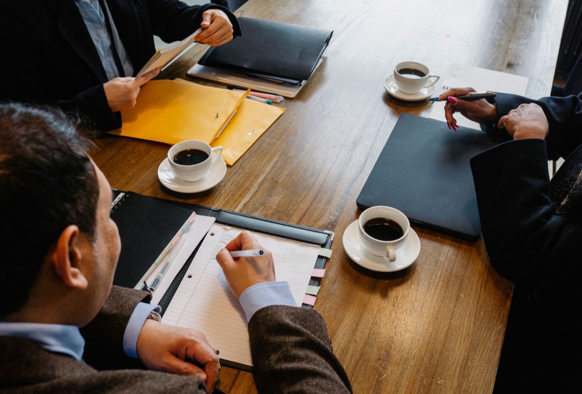 People seated around a wooden meeting table with coffee cups, folders, an envelope and a notepad