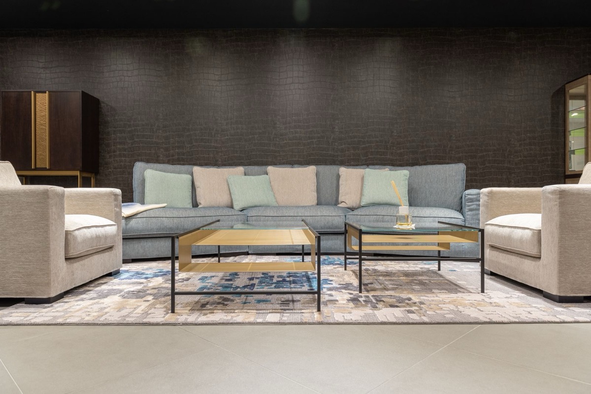 A lounge with a grey sofa, two beige armchairs and glass coffee tables on a patterned rug