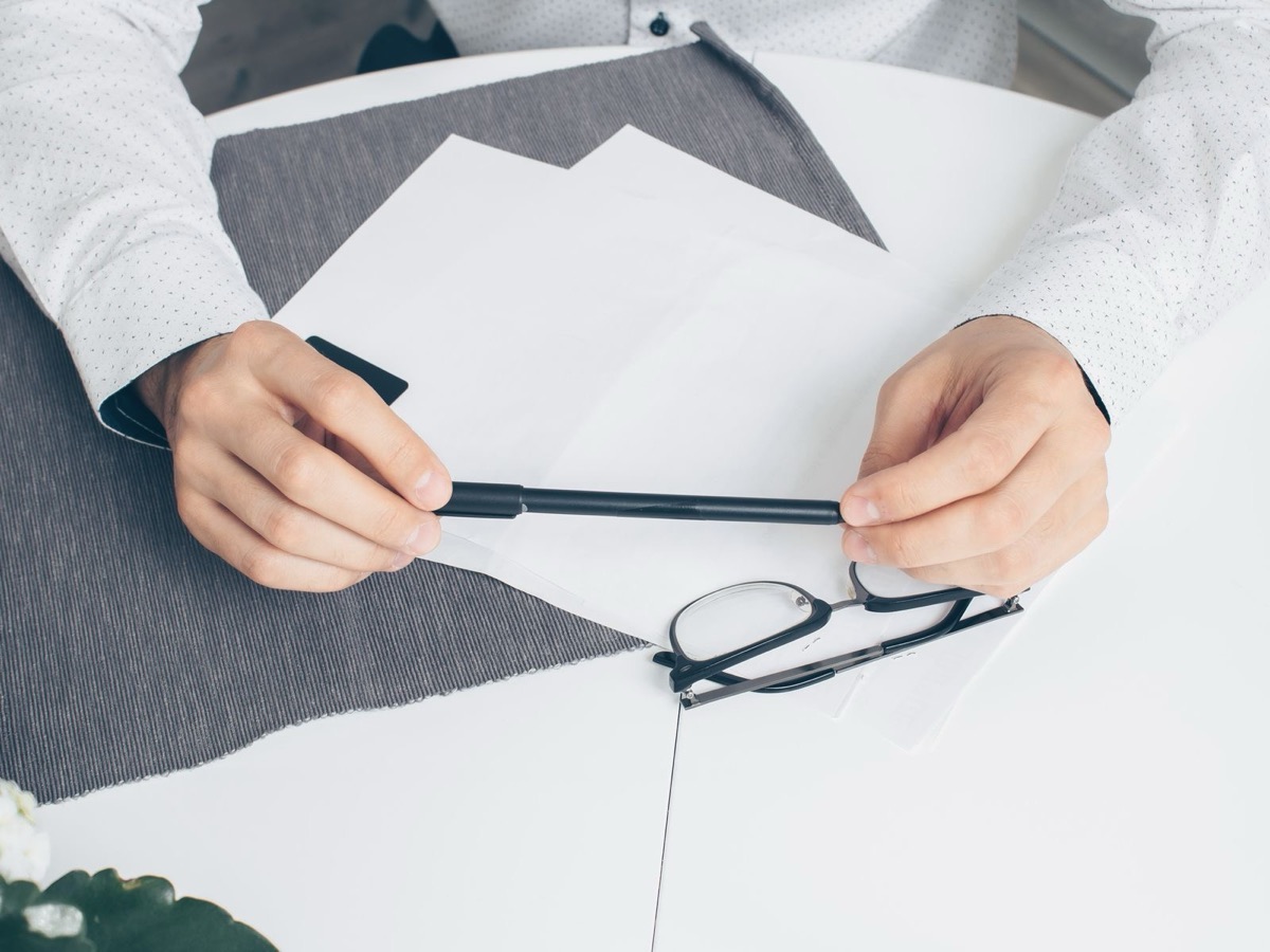 Hands holding a plain black pen above blank white sheets on a white table beside a pair of glasses