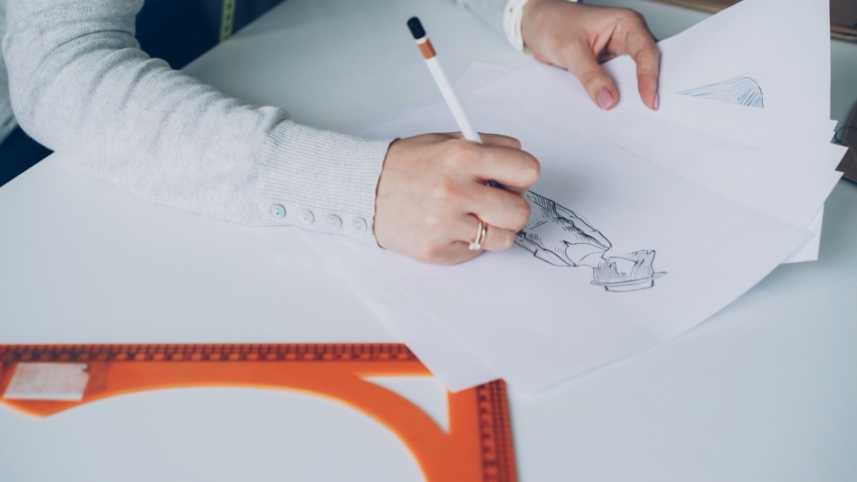 A hand sketching on sheets of paper at a white desk beside an orange ruler