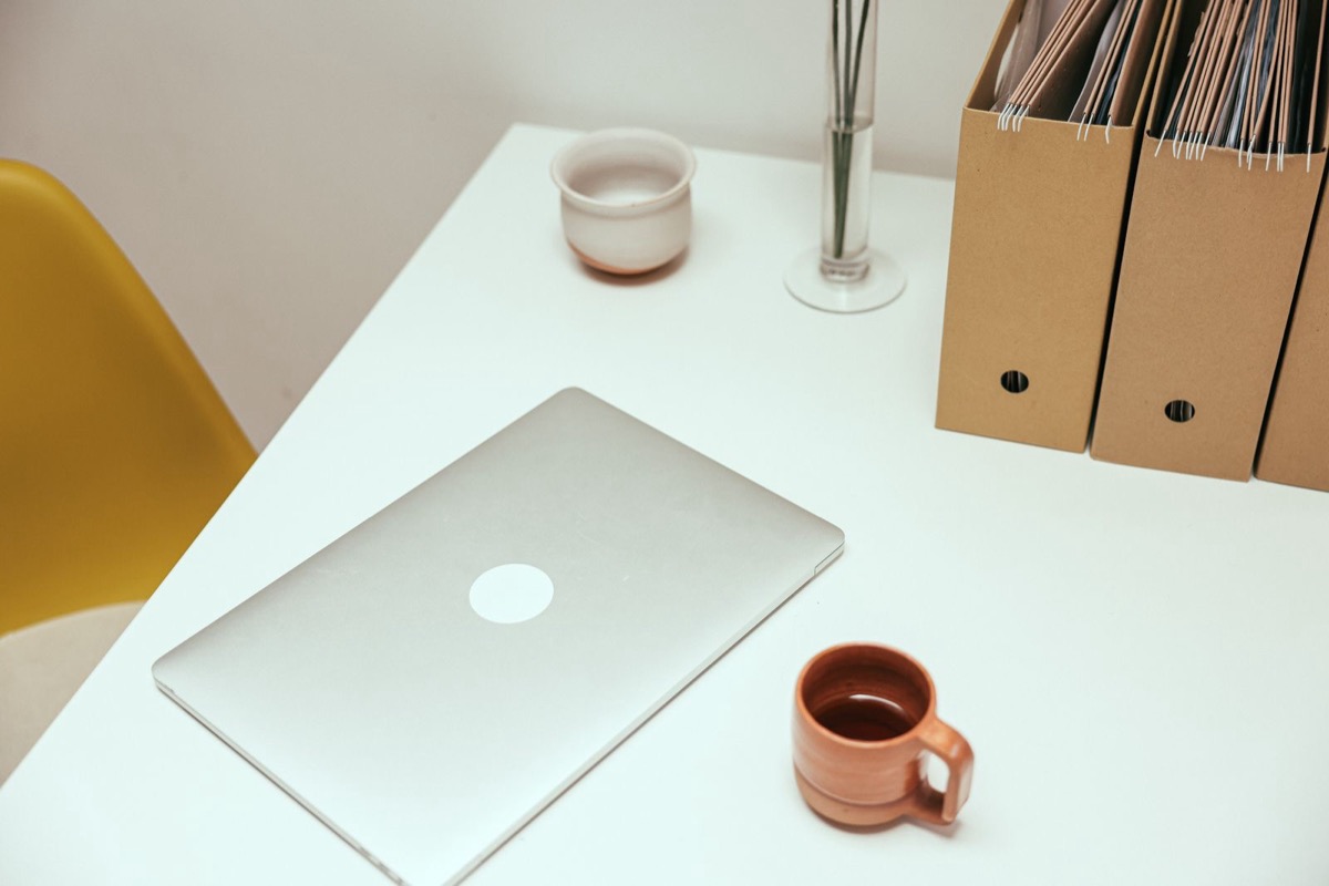A closed laptop, a mug and cardboard file boxes on a white desk beside a yellow chair