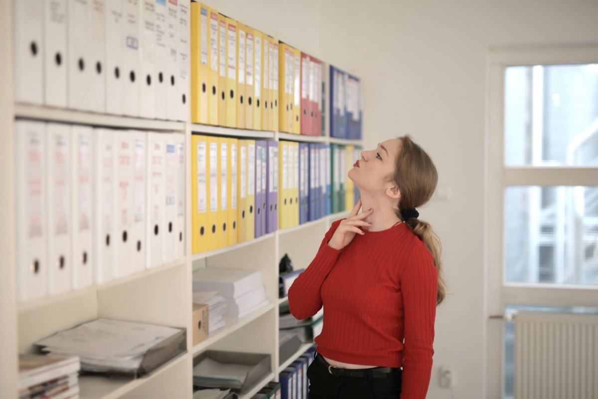 A woman in a red jumper looking up at shelves of ring binders in a bright office