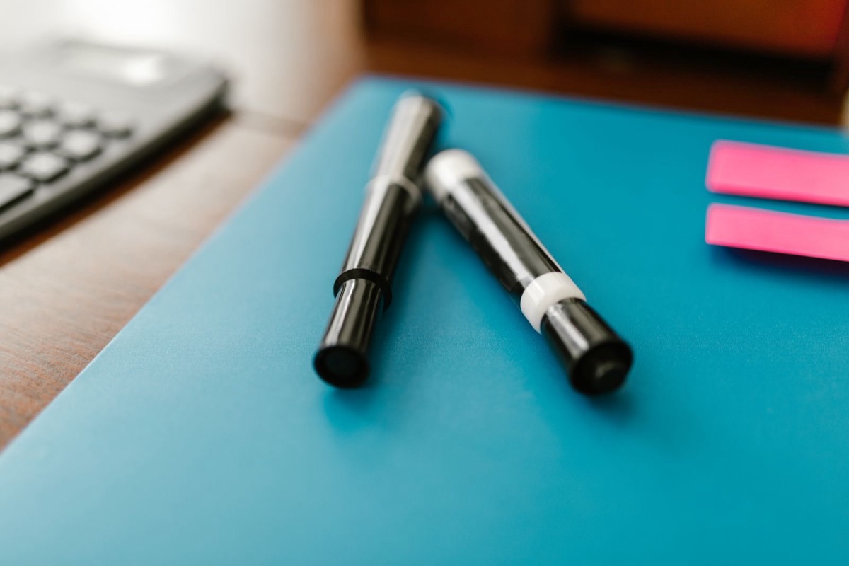 Two black marker pens on a blue folder, with a calculator and pink sticky notes on a wooden desk